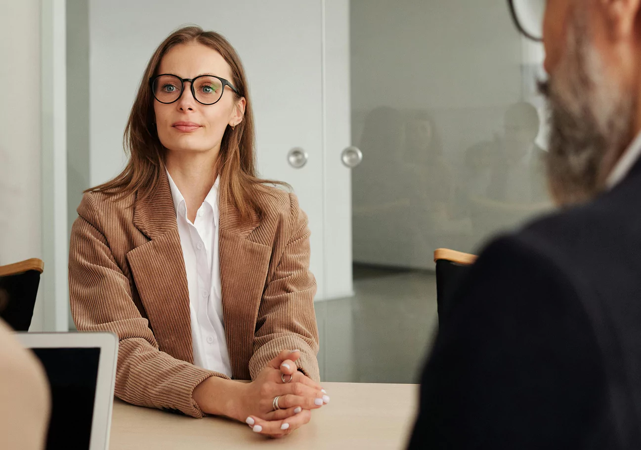 A woman wearing glasses, a white blouse, and a tan corduroy blazer sits at a table with her hands clasped during a professional meeting
