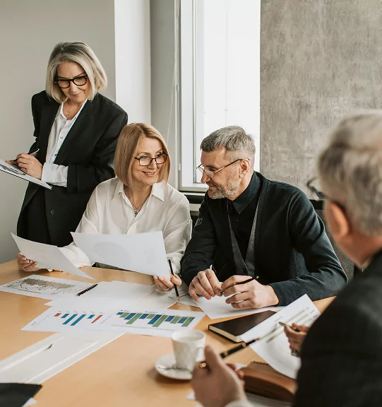 A group of four professionals, two men and two women, are gathered around a conference table reviewing printed charts and documents.